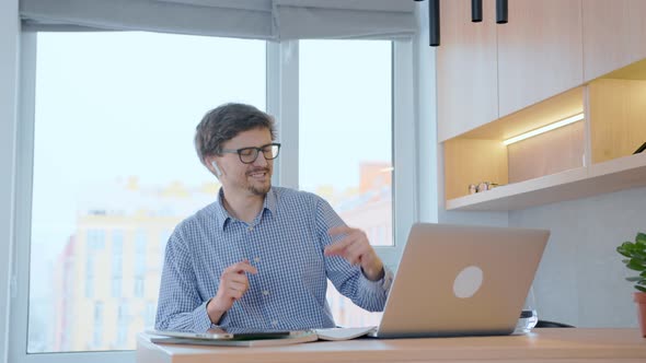 Happy young man listens to music on headphones while sitting at his desk, smiling and dance. alt