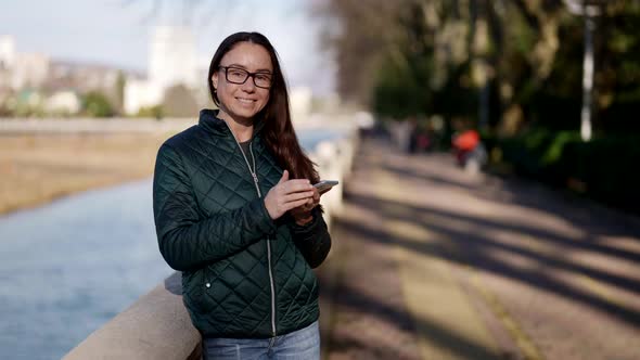 Portrait of a Beautiful Adult Woman with Glasses Standing on the Street in Spring with a Phone in alt