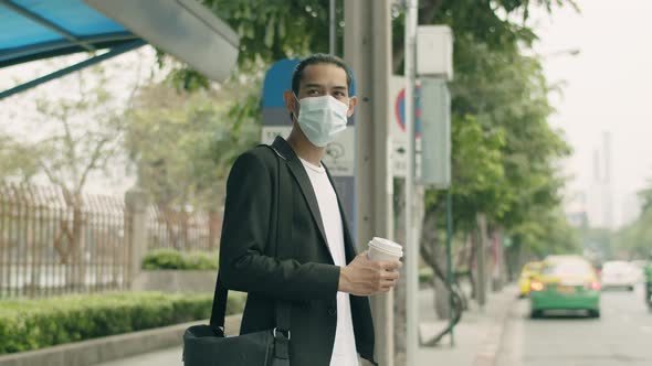 Asian businessman wearing a face mask holding coffee cup standing at a bus stop.