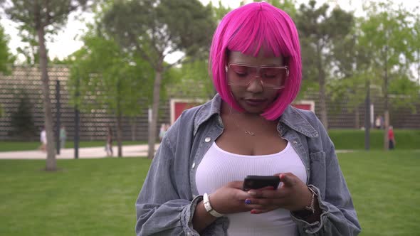 A Young Stylish Black Woman with Pink Hair Uses Her Smartphone Outdoors alt