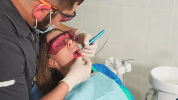 Doctor Orthodontist with Glasses Whitens the Teeth of a Young Woman with a Laser alt