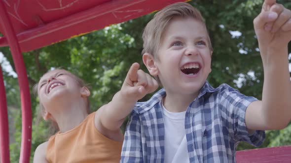 Cute Boy and Girl Pointing Away with Fingers Sitting on the Swing Close Up in Park, Laughing Happily alt