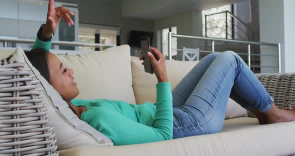 Mixed race woman lying on couch having video chat on smartphone blowing kisses alt