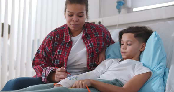 Black Mother Visiting Son Lying with Broken Arm in Bed in Hospital Ward alt