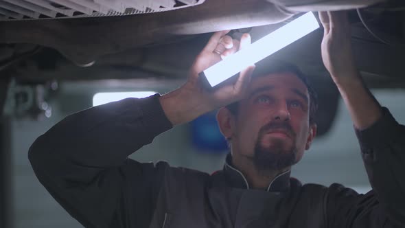 Portrait of the Mechanic Inspecting the Car From Below Raised on the Lift and Inspecting the Bottom alt