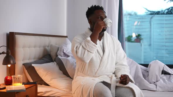 Confident Young Handsome Man Drinking Coffee Looking Away Smiling Sitting on Bed Indoors alt