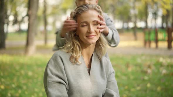 Closeup of Relaxed Beautiful Slim Woman Sitting in Autumn Park As Joyful Boy Running From Background alt