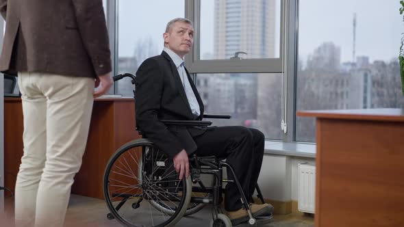 Side View of Sad Disabled Senior Man in Wheelchair Looking Out the Window in Office and Talking to alt