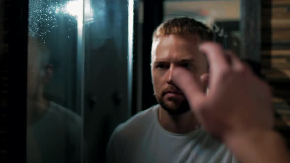 Man wipes a misted mirror with his hand and look at his reflection in the bathroom alt