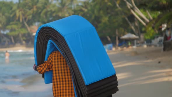 Dark Skinned Man Holds Mats on Head and Walks Along Beach alt