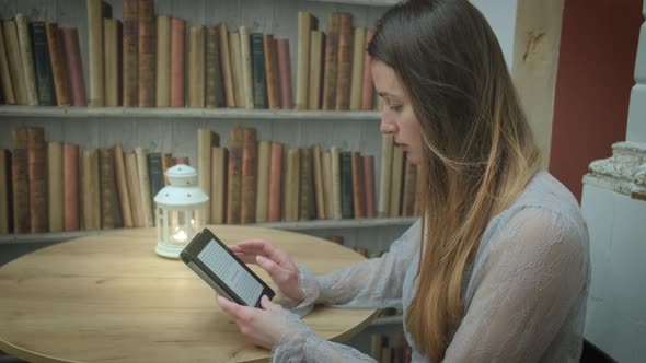 Close Up of Attractive Caucasian Woman Reading Ebook at Table with Candle of Bookshelf alt
