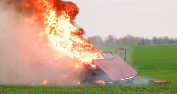 A red car explodes in an open field. The car is on fire.