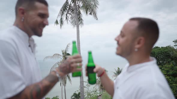 Male Gay Couple Clinking Bottles of Beer and Smiling While Resting on Tropical Island Standing at alt