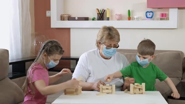 Grandma Plays with Children in Medical Masks with Wooden Blocks in the Room. Social Distancing and