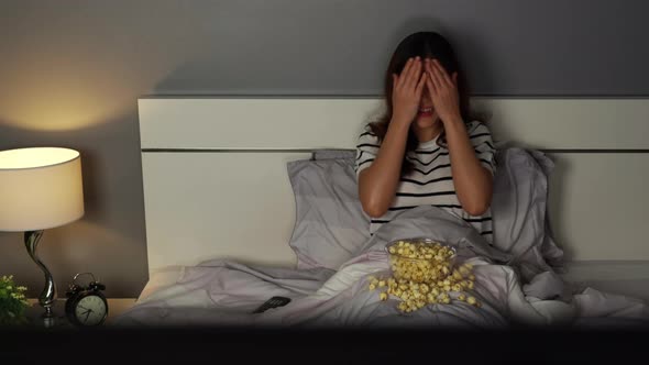 shocked young woman watching horror movie TV on a bed at night