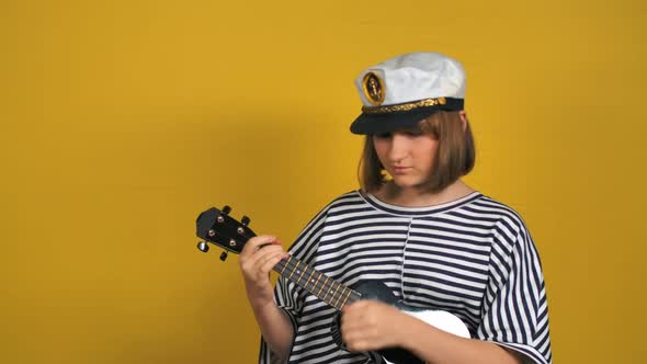 Young Beautiful Blonde Girl in Sailor Hat Plays the Ukulele Guitar in the Studio alt