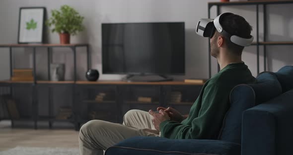 Male Gamer Is Playing Online with Headmounted Display and Joystick Sitting Alone in Living Room on alt