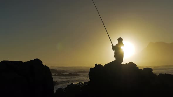 Fisherman on rocks reeling in fish during  golden sunset over ocean, silhouette against sky. alt