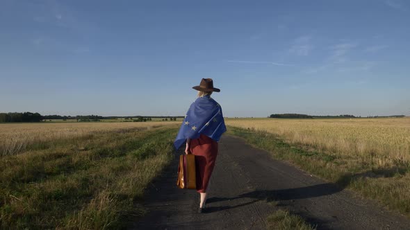 Girl in EU flag with suitcase on country road in sunset