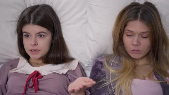 Two Confident Young Women Discussing Rumors Lying on Comfortable Soft Pillow Indoors alt