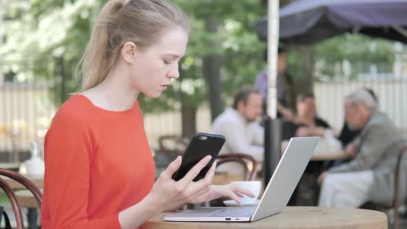 Woman Using Smartphone and Laptop Sitting in Outdoor Cafe alt