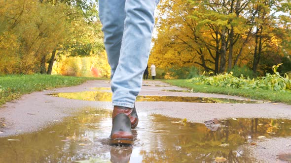 Woman in Boots Walks Along Road with Puddles in Autumn Park alt
