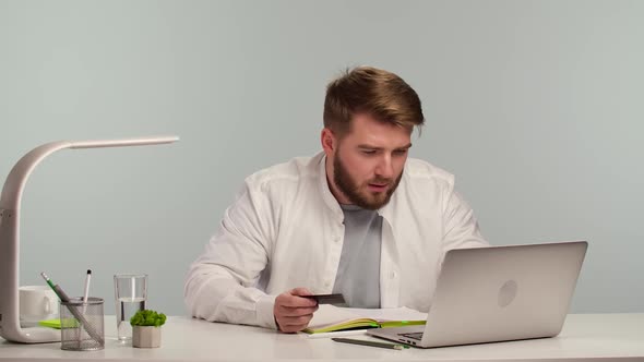Young Man Sits at Laptop in His Home Office and Makes Online Shopping Using Bank Card alt
