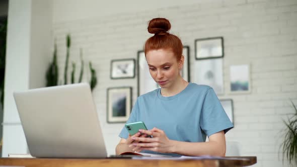 Attractive Redhead Woman Is Using Mobile Phone While Sitting at Desk with Laptop alt