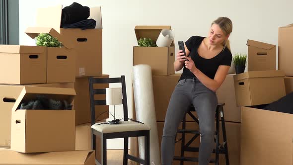 A Moving Woman Sits on a Chair in an Empty Apartment and Takes Pictures of a Lamp on Another Chair alt