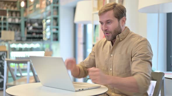 Young Man Celebrating Success on Laptop in Cafe alt