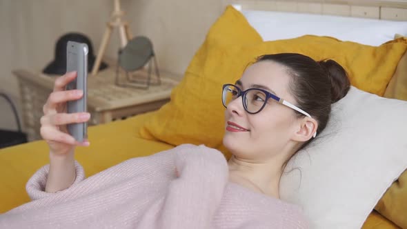 Woman waving during a smart phone video call lying on a bed at home