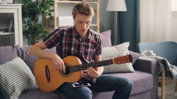 Young Musician Is Tuning Acoustic Guitar Touching Strings Sitting on Couch at Home During Leisure alt