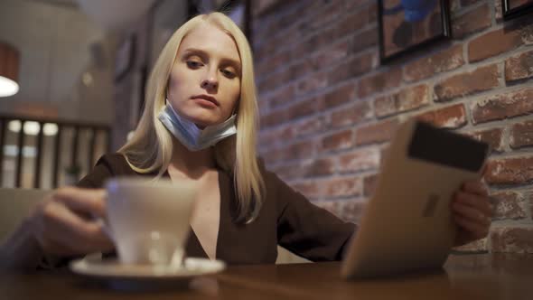 A Young Woman in a Medical Mask Drinks Delicious Coffee in a Restaurant and Scrolls the Tablet