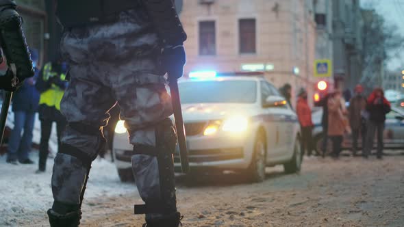 Armored Policeman with Batons Stands in Front of Car with Flashing Strobe Lights alt