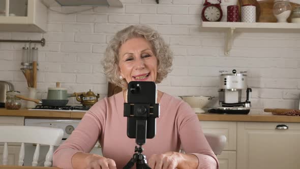 Pensioner Blogger Speaks at Smartphone on Tripod in Kitchen alt
