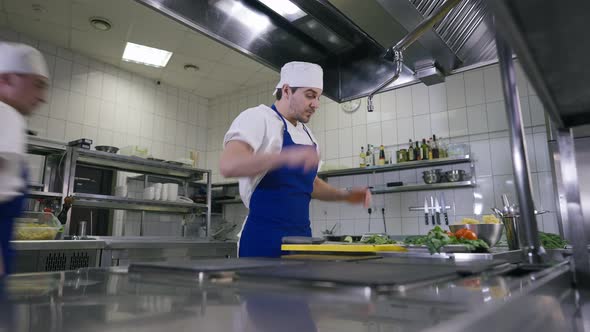 Tired Young Handsome Cook Standing in Kitchen Wiping Forehead As Colleague Entering alt
