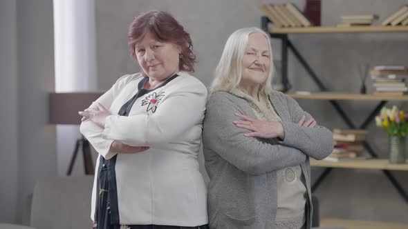 Confident Senior Caucasian Women Crossing Hands Looking at Camera Standing Back to Back Indoors alt