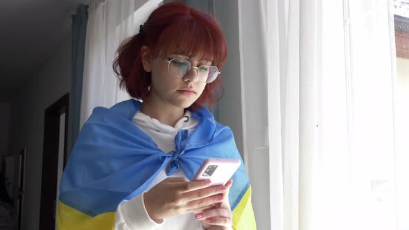 Young redhead ukrainian teen girl with flag and mobile phone near window