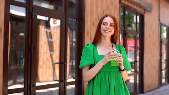 Portrait of Smiling Attractive Young Woman Drinking Cocktail Through Straw Standing at City Street alt
