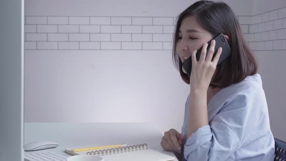 Asian woman working on laptop while sitting in a living room using phone for work in her home alt