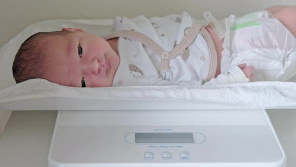 Weighing newborn baby in white clothes on a home scale alt