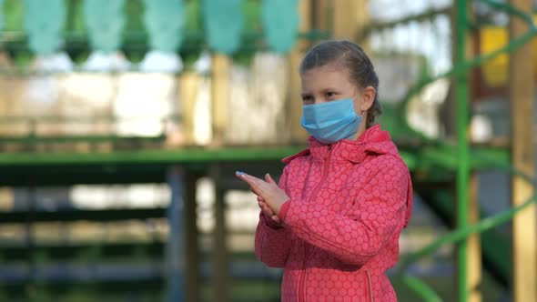 A girl in a medical mask. Her mother sprays disinfectant on her hands in the Playground.