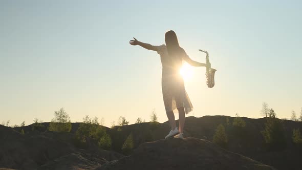 Slender Woman in White Dress Stands on Top of Mountain Hands to the Side Holding Saxophone in One alt