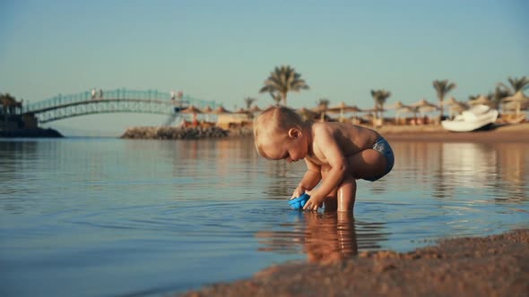 Funny Baby Boy Pouring Water Into Blue at Calm Sea Bay in Evening Time. alt