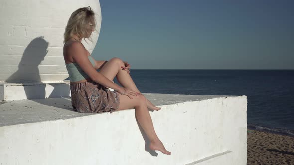 An Attractive Woman in a Summer Dress is Sitting on the Embankment Against the Backdrop of the Sea alt