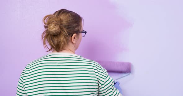 Young Tired Woman Listlessly Paints Wall with Paint Roller in Lilac Color