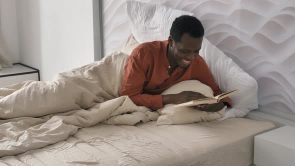 Happy AfricanAmerican Man in Pajama Reads Book Lying in Bed alt