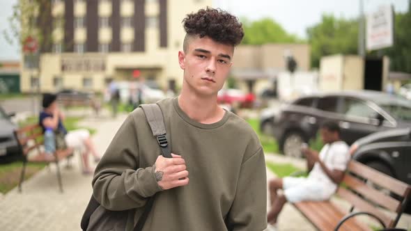 A young man with a backpack looks at the camera, standing on the city alley alt