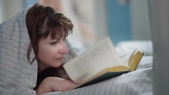 Woman Lies on the Bed Near the Window and Reads a Book Reads with Pleasure