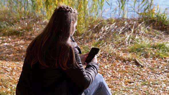 Young Woman Reading Text Message in a Smartphone Sitting on Fallen Dry Leaves alt